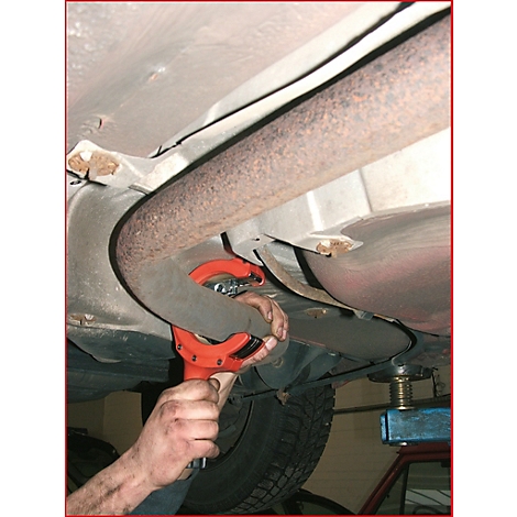 Ein Mechaniker schneidet ein Auspuffrohr mit einem roten Werkzeug unter einem Auto ab.